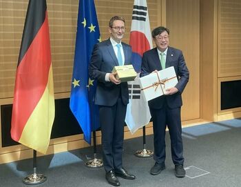 Stefan Schnorr und Yun Kyu Park halten Geschenke in der Hand und stehen vor der deutschen, europäischen und südkoreanischen Flagge.