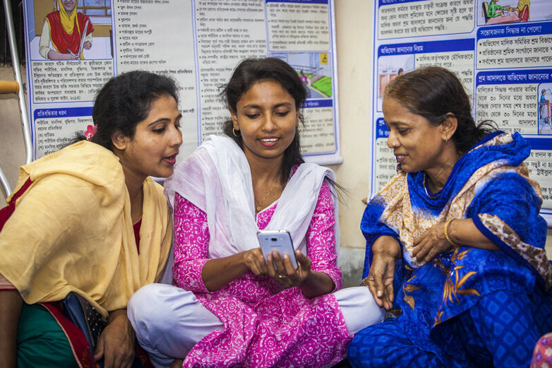 Drei bengalische Frauen in bunter Kleindung betrachten etwas auf einem Mobiltelefon. Eine von ihnen hat eine Behinderung an den Armen.