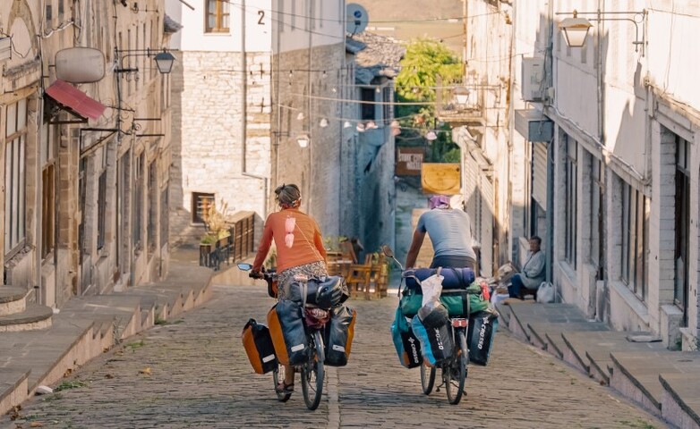 Zwei Personen fahren auf schwer beladenen Fahrrädern eine Straße mit Kopfsteinpflaster hinunter.