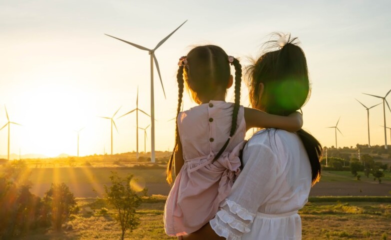 Eine Frau mit einem kleinen Mädchen auf dem Arm vor Windrädern und einem Sonnenuntergang. Das Bild ist in Blickrichtung der Frau aufgenommen.