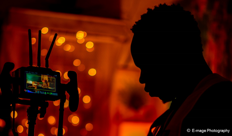 Die Silhouette einer Person, die vor einer Kamera steht, ist vor einem rötlichen Hintergrund mit warmen Lichtern erkennbar.