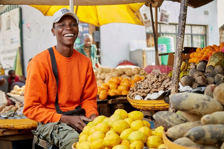 Eine lachende Person sitzt an einem Marktstand voller Früchte.