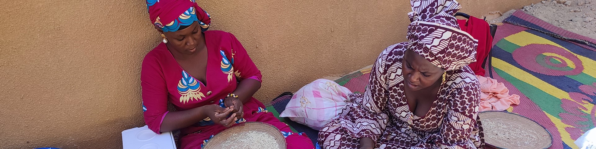 Zwei Frauen der Frauenvereinigung in Dirè sitzen mit großen Tellern im Schneidersitz auf dem Boden und verarbeiten Weizen.