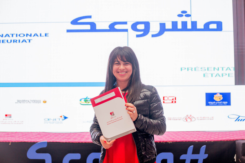 Eine junge Unternehmerin steht lachend auf einer Bühne und hält eine Auszeichnung des nationalen Unternehmertumwettbewerbs in den Händen.