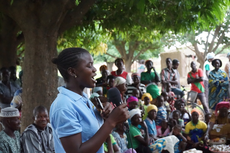 Eine junge Frau hält ein Mikrofon in der Hand und spricht vor einer Menschengruppe über das Thema weibliche Genitalverstümmelung.