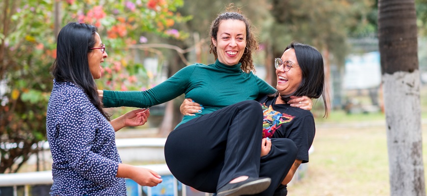 Drei Frauen stehen in einem Kreis beisammen in einem Park. Die Frau rechts hat eine andere Frau auf dem Arm. 