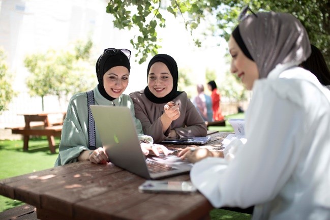 Drei Frauen mit Kopftüchern sitzen an einem Tisch und arbeiten an einem Laptop.