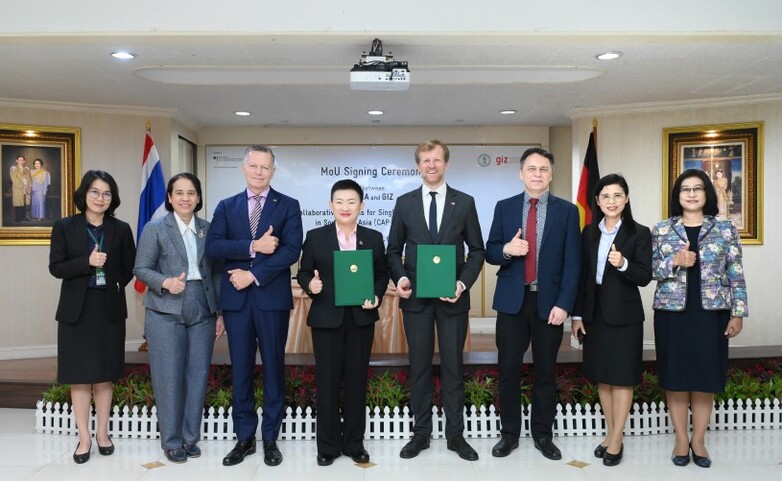 Vertreter*innen aus Deutschland und Thailand halten bei einer Unterzeichnungszeremonie Dokumente, während andere Personen aus beiden Ländern Daumen hoch zeigen.
