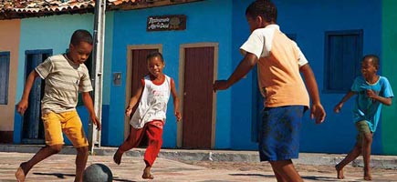 Four children play soccer in front of colourful houses.