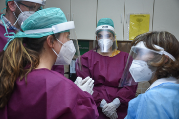 Three women wearing protective medical attire engage in conversation.