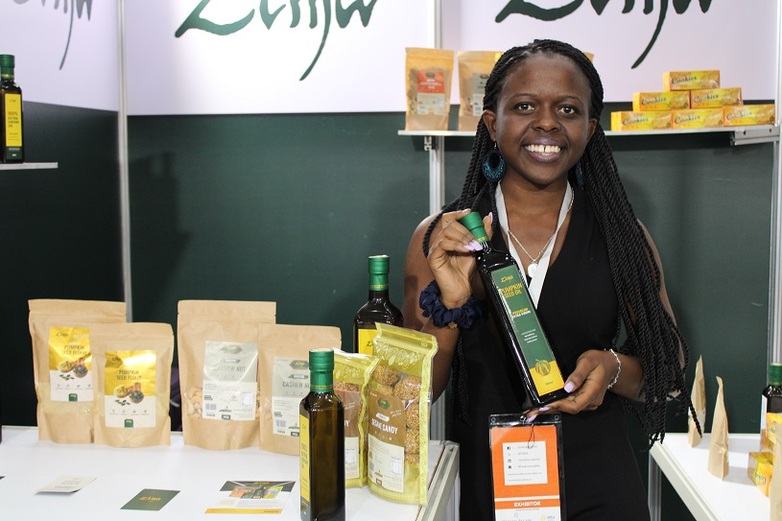 A woman stands at a market stall and smiles as she holds a bottle of pumpkin seed oil up to the camera.