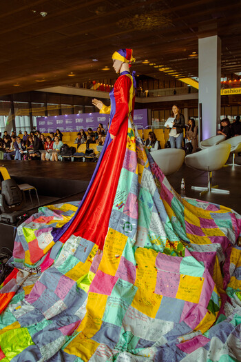A doll with a dress made of patches can be seen in the foreground. An event is being held in the background.