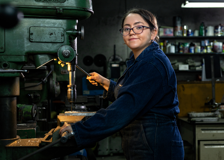 A young woman in blue work clothing stands in front of a bench drill, facing the camera. © GIZ / Photographer: Carlos Agüero