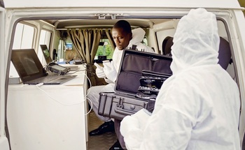 Two men in protective gear are working in a van a computers.