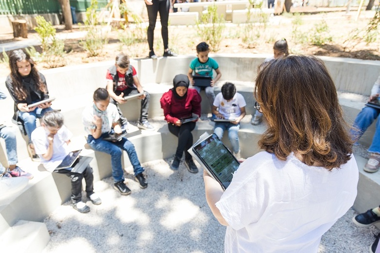 Schoolchildren sit on round benches opposite their teacher in an open-air classroom, holding tablets in their hands.