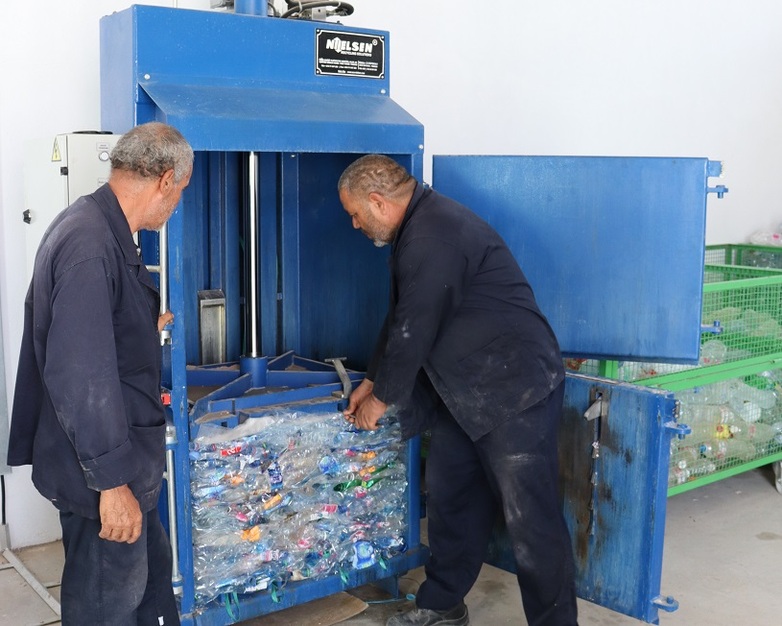 Two men in work clothes operating a baler machine in a waste sorting plant.
