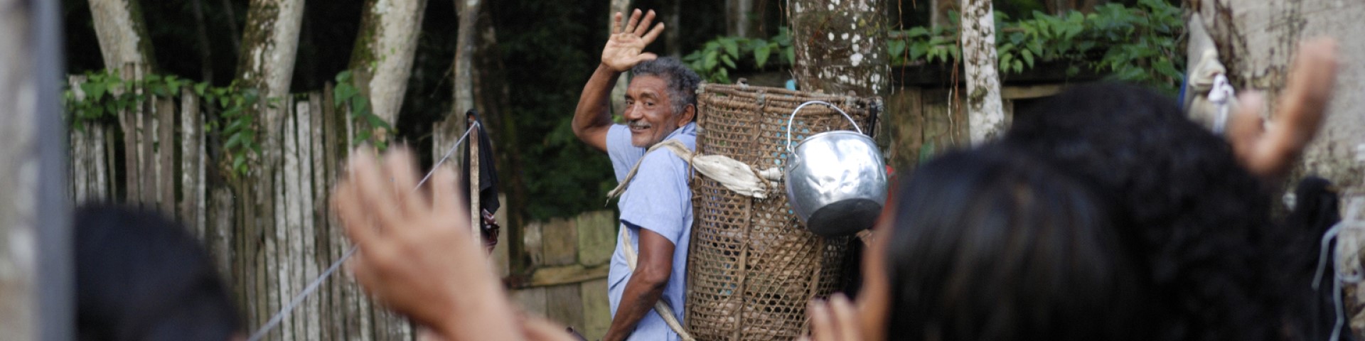 A man carrying a basket on his back stands in front of a forest and waves to a group.