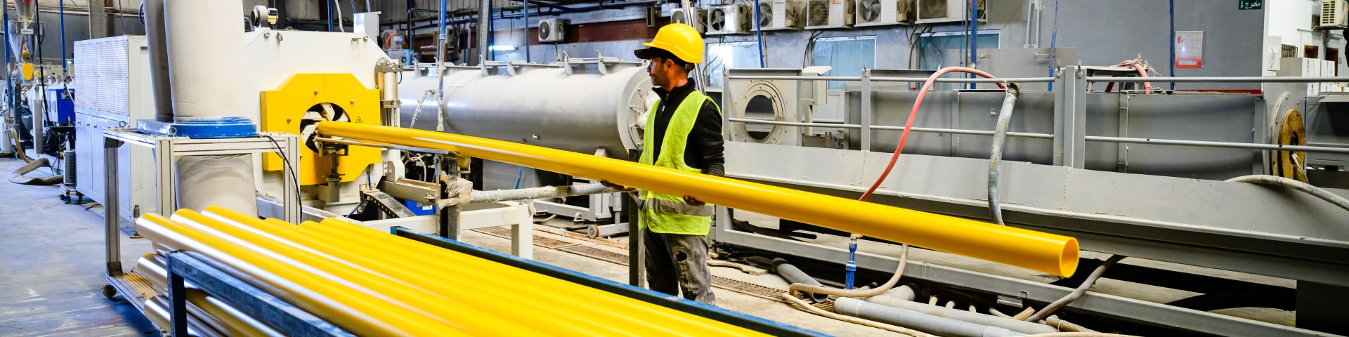 A worker operates machinery in a factory, handling a large yellow plastic pipe.