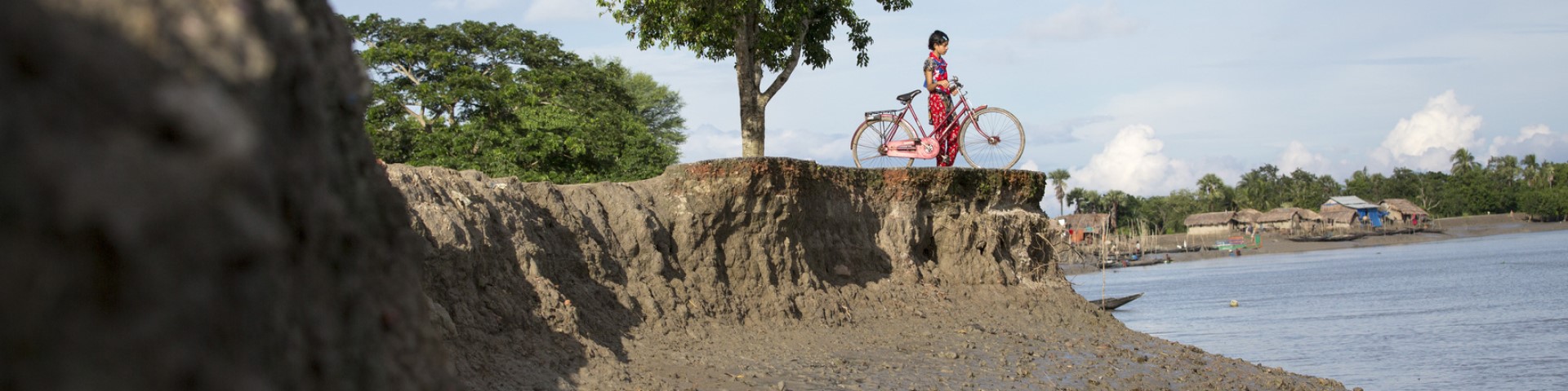 A person stands with their bicycle on a riverbank that is affected by erosion.