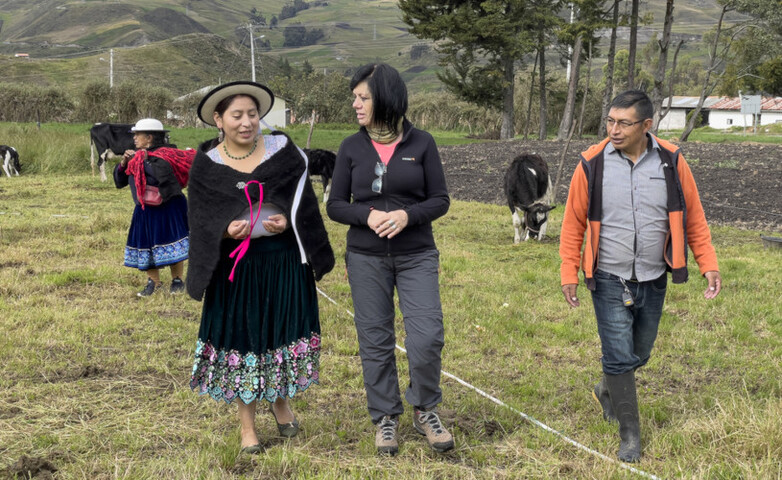 Three people, one of them in traditional costume, in conversation beside a field and a cow pasture.
