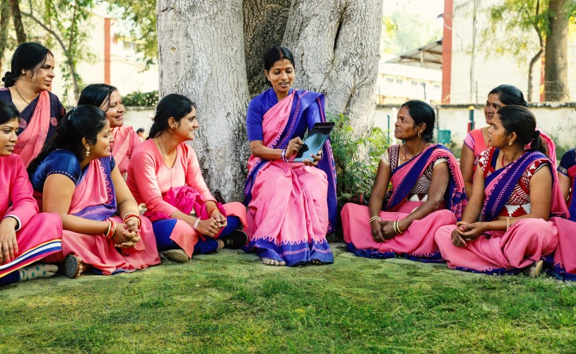 A group of women are sitting in a circle. One of the women holds a tablet in her hand and explains to the other women what is on it.