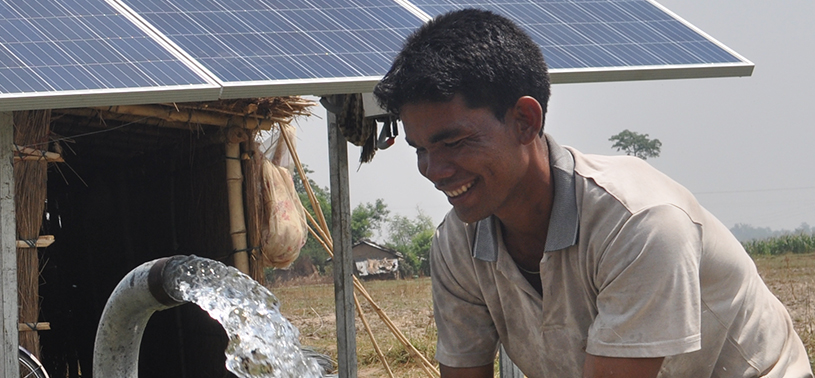 A gush of water from a pipe and a smiling man in front of a hut with solar panels on the roof.