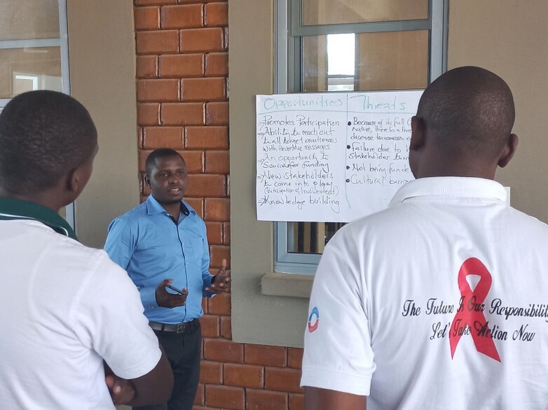 A person speaks during a professional development course for DACAs and PACAs in Chongwe, standing next to a poster while two other people listen.