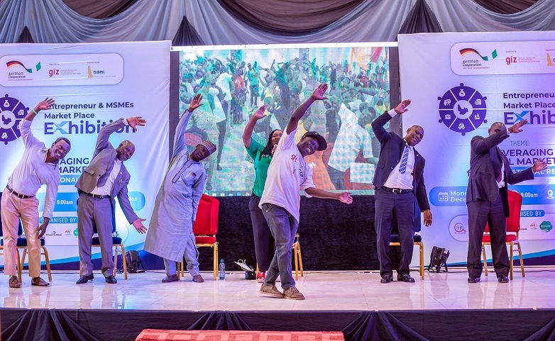 Seven people stand on a stage at a conference on entrepreneurship, raising their arms in the air with a smile.