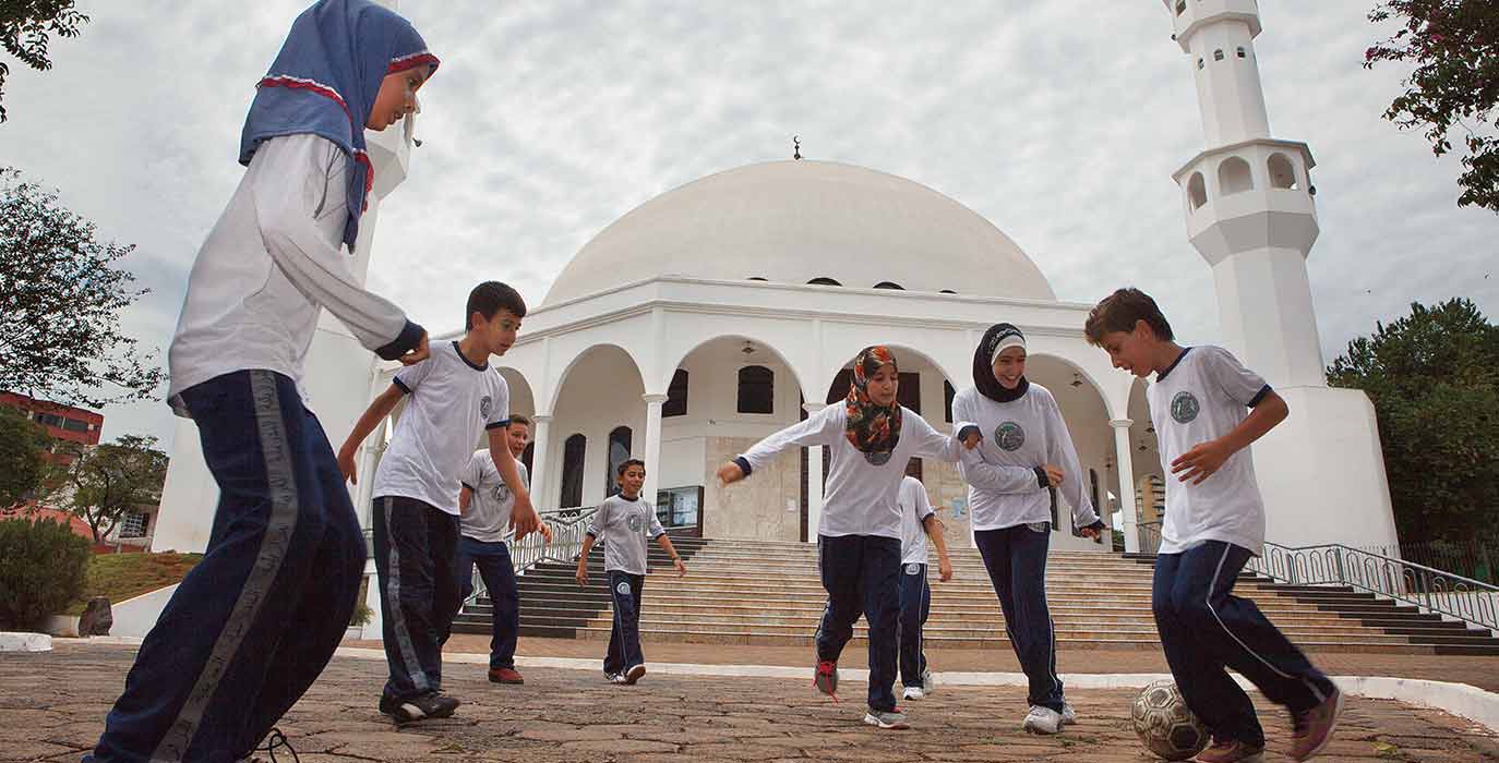 Girls with headscarfs and boys playing football, mosque in background