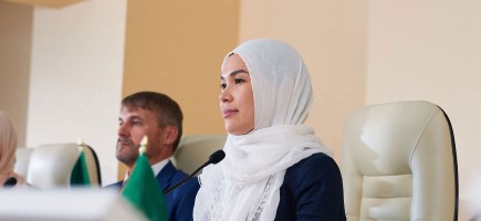 A person wearing a hijab sits at a conference table with microphones and flags in front of her.