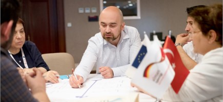 A man draws something on a document. Other people are sitting around him and there are small flags on the table, including those of the EU and Turkey.
