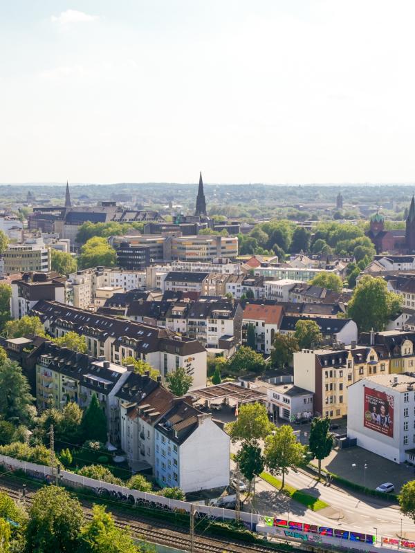 Luftaufnahme einer Stadt mit dicht bebauten Wohnhäusern, mehreren Kirchtürmen und Bahngeleisen im Vordergrund, umgeben von vielen Bäumen und Grünflächen.
