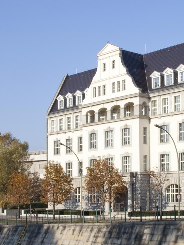 Historisches, mehrstöckiges Gebäude mit dunklem Schrägdach und zahlreichen Fenstern am Ufer eines Kanals; umgeben von Bäumen und modernen Gebäuden im Hintergrund, bei klarem Himmel.