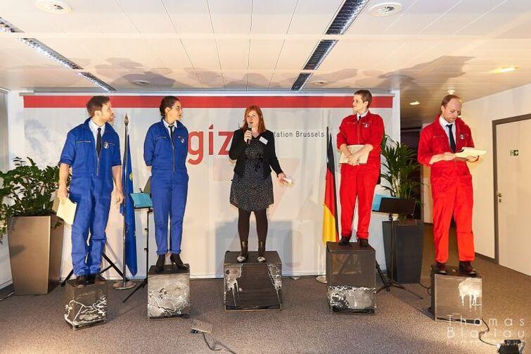 5 Personen stehen auf Podesten. Zwei in blau eine in schwarz und zwei in rot