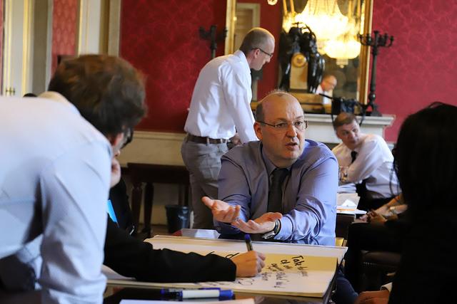 Ein Mann sitzt vor einem Plakat und redet zu seinen Sitznachbarn