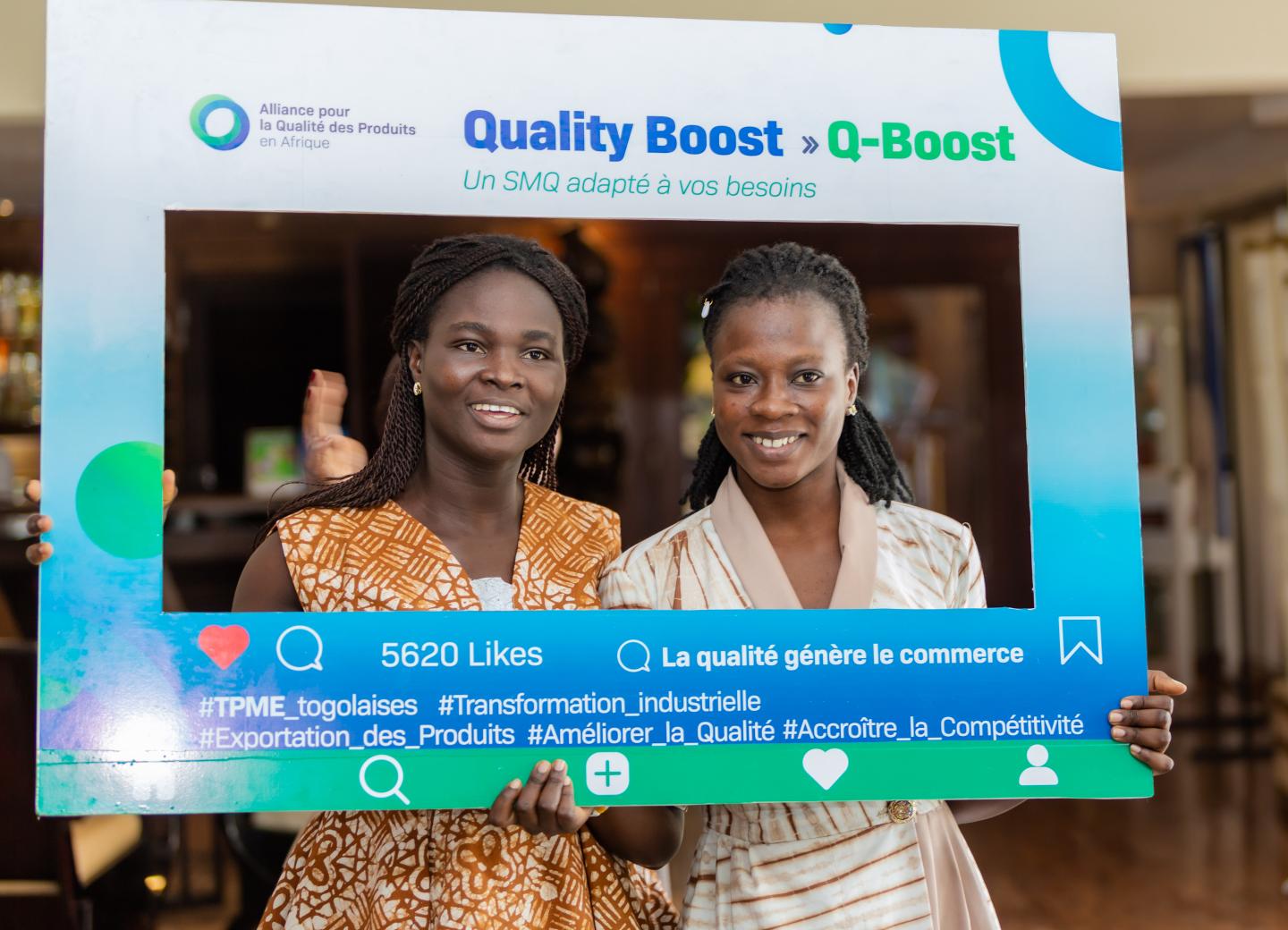 Two women smile while posing with a “Quality Boost (Q-Boost)” promotional frame.