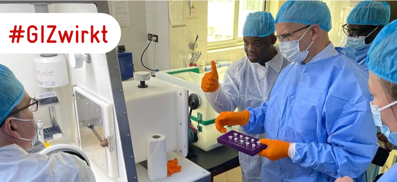 Fünf Personen in medizinischer Schutzkleidung und Maske stehen um ein Laborgerät herum.