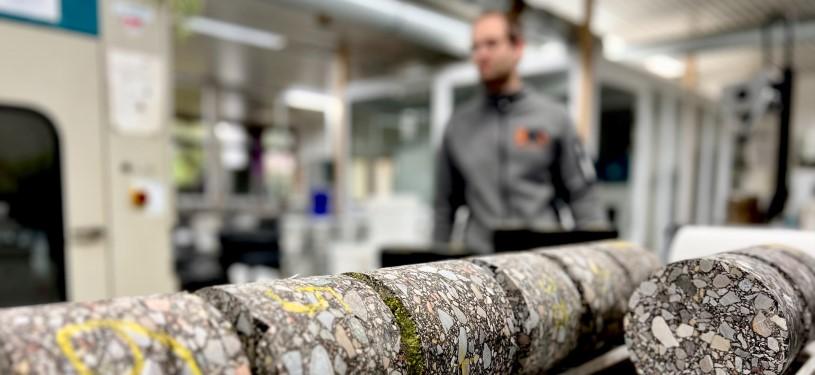 Stücke eines nachhaltigen Straßenbelags im Querschnitt, im Hintergrund eine Fabrik von innen.