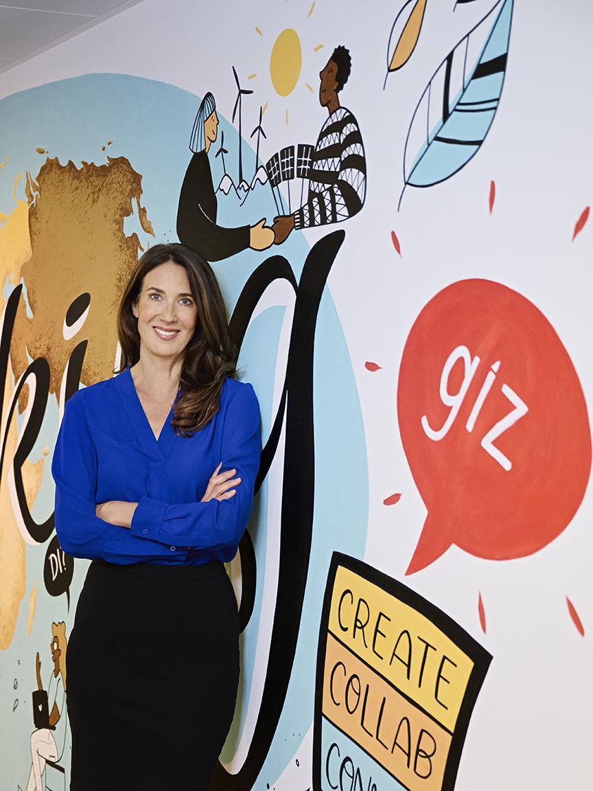 A woman with long brown hair wearing a blue blouse sits at a conference table in a modern office with glass walls and round ceiling lights in the background.