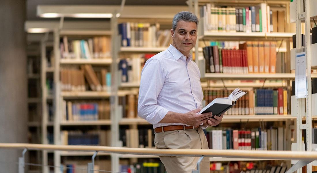 A man stands in a library with an open book in his hand.