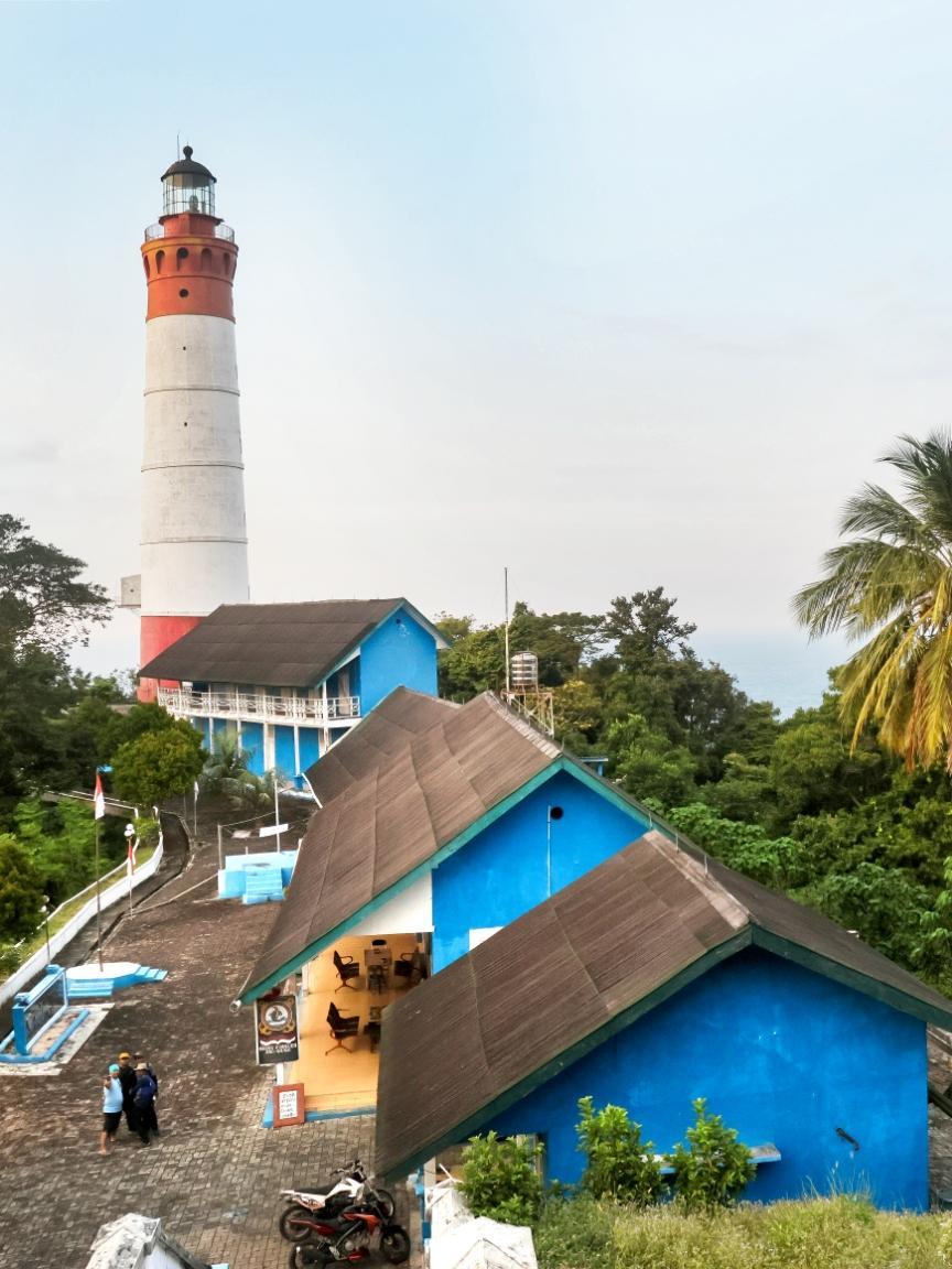 Ein rot-weißer Leuchtturm ragt über mehrere strahlend blaue Gebäude mit schrägen Dächern empor, umgeben von tropischer Vegetation.