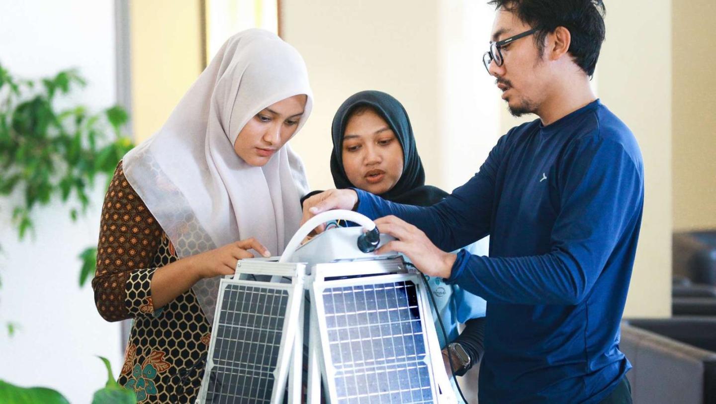 Zwei junge Frauen mit Hijab und ein Mann untersuchen gemeinsam ein technisches Gerät mit Solarpanels in einem Innenraum.