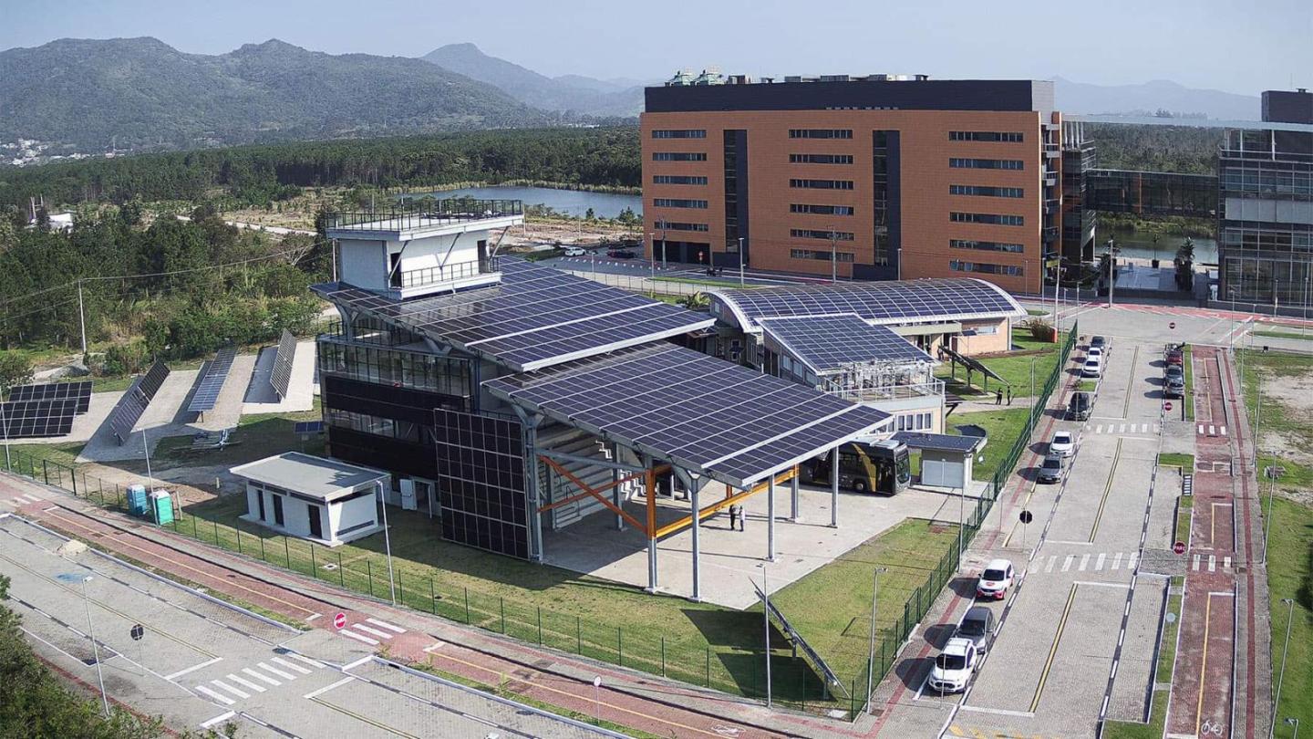 A modern building with numerous solar panels on the roof and surrounding fields, set in green countryside with mountains in the background.