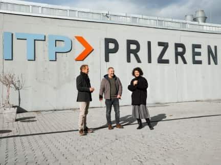 Three people are standing in front of a grey wall with the words ‘ITP PRIZREN’ written on it, chatting.
