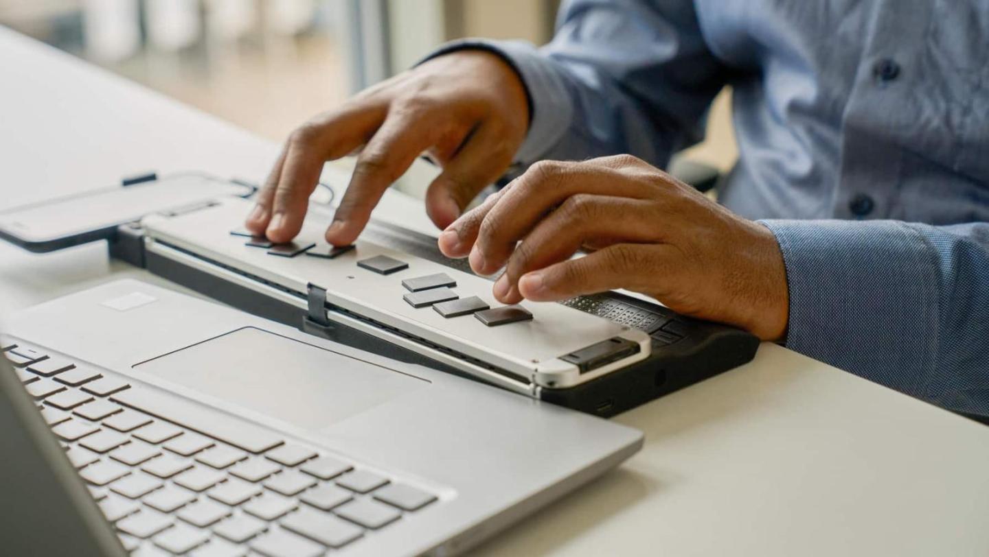 Close-up of hands typing on a Braille display in front of a laptop.