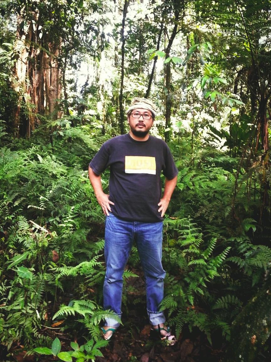 A man wearing glasses, a black T-shirt, jeans and sandals stands in the middle of a dense tropical rainforest with lush ferns and tall trees.