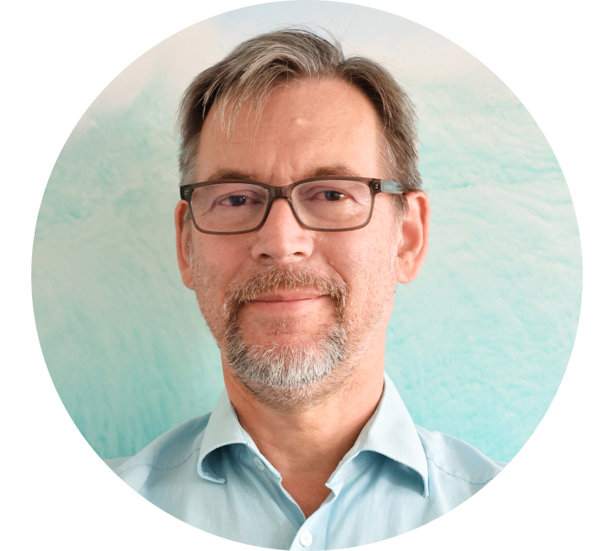 Porträt eines älteren Mannes mit Brille, grauem Bart und hellblauem Hemd vor einem pastellfarbenen Hintergrund.