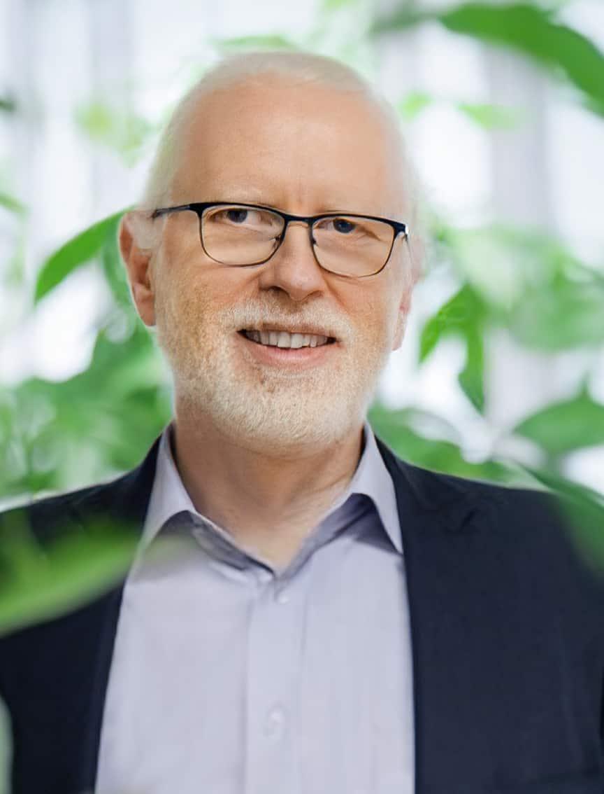 Portrait of an elderly man with a white beard, glasses and a dark jacket, surrounded by green plants.