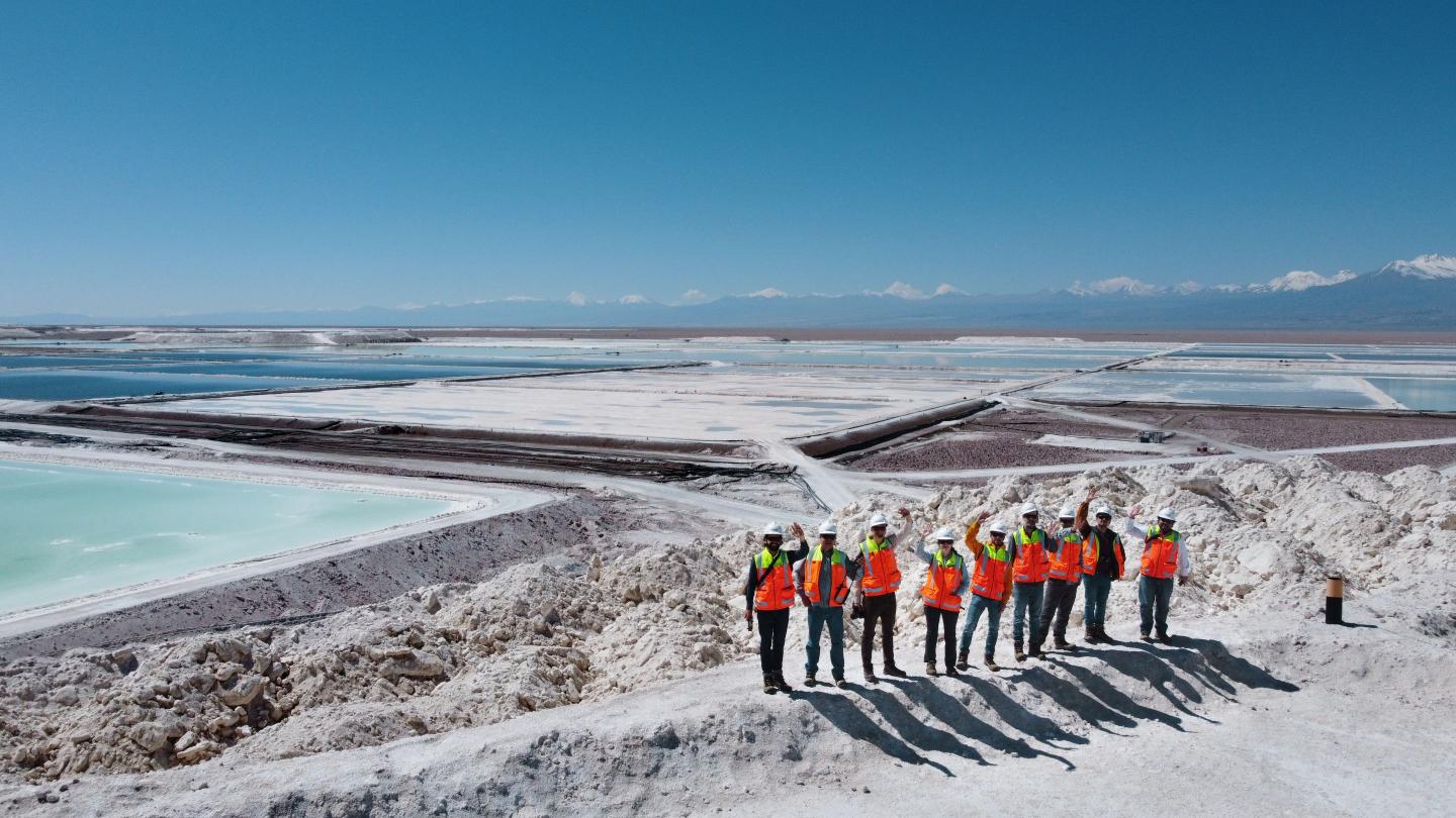 Gruppe von Personen mit Sicherheitshelmen und leuchtend orangen Warnwesten steht auf einem Hügel in einem Salzabbaugebiet mit grossen Verdunstungsbecken; im Hintergrund sind Berge und ein klarer blauer Himmel sichtbar.