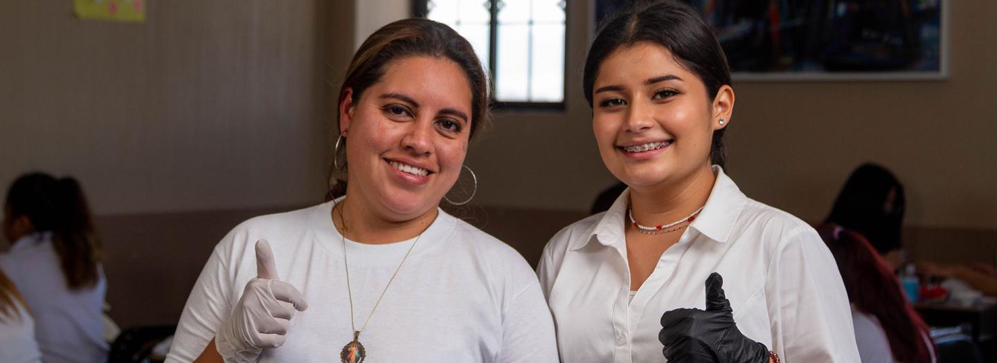 Drei Jugendliche in Arbeitskleidung mit Hörschutz strecken ihren rechten Daumen hoch und schauen in die Kamera. Vor ihnen stehen auf einem Arbeitstisch Holzprodukte wie gedrechselte Möbelbeine.